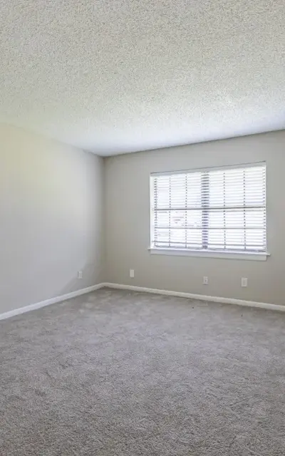 A vacant room with light beige walls and gray carpet, featuring a window with wooden blinds.