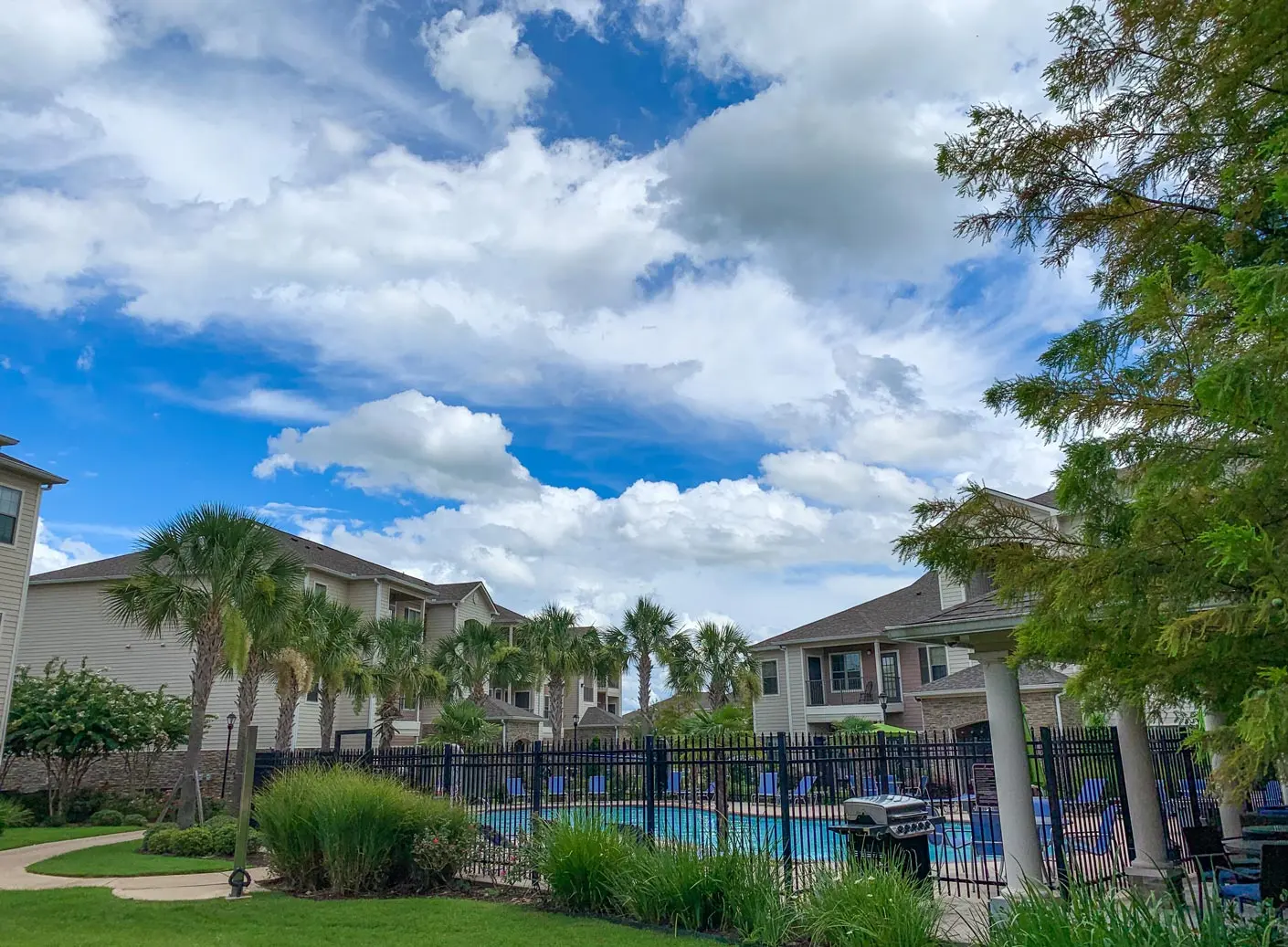 A tranquil poolside scene featuring a well-maintained pool surrounded by palm trees and apartment buildings under a partly cloudy sky.