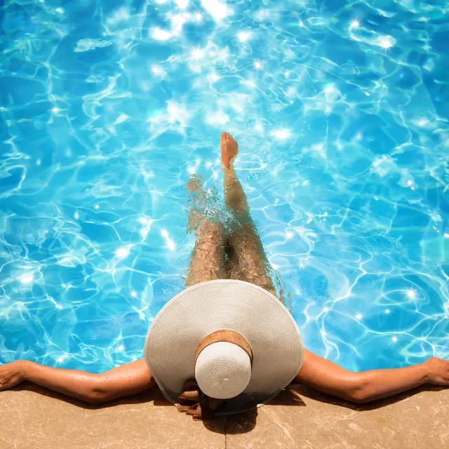 Person relaxing by a swimming pool, wearing a wide-brimmed hat, with arms outstretched and feet in the water.