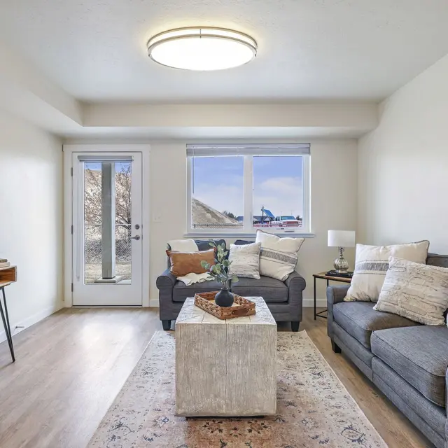 Bright and Cozy Living Room A cozy living room featuring a gray sofa with assorted pillows, a light-colored area rug, and a coffee table with decor. There is a mirror on the wall and a console table with flowers. A glass sliding door leads to an outdoor view.