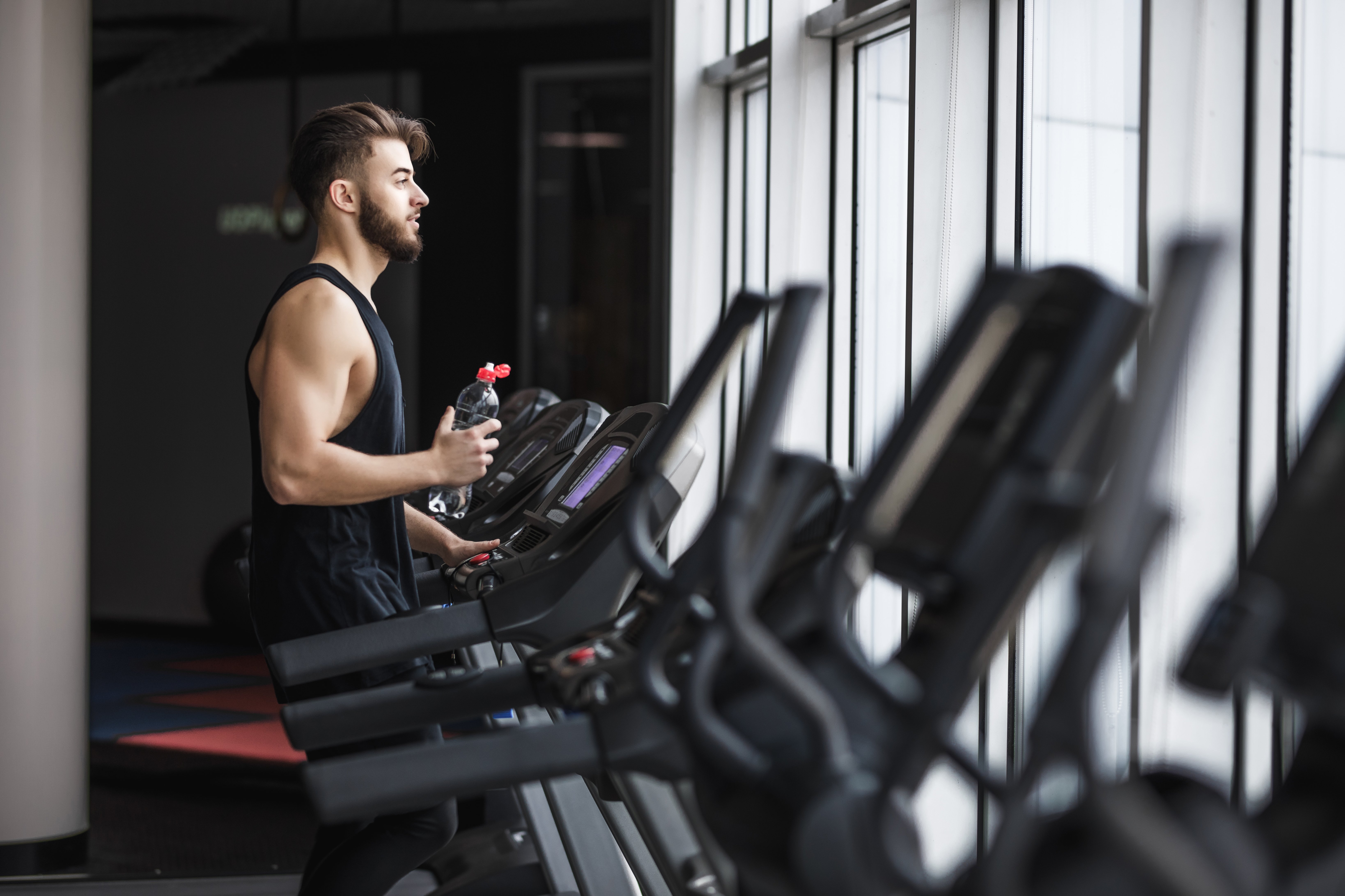 A young man in a sleeveless black shirt is exercising at a gym. He stands on an elliptical machine while holding a water bottle, looking out of large windows that let in natural light.