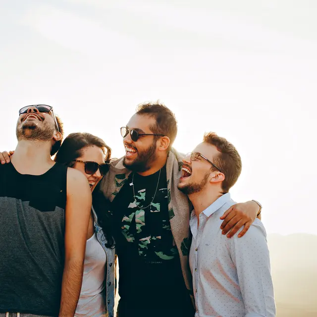 Group of four friends laughing together outdoors during sunset, wearing sunglasses and casual clothing, with rolling hills in the background.