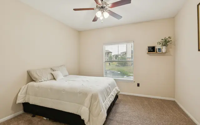 Cozy Bedroom Interior A cozy bedroom featuring a bed with beige bedding, a ceiling fan, and a window with natural light. The walls are painted in a light color and a small shelf with a plant is visible next to the window.