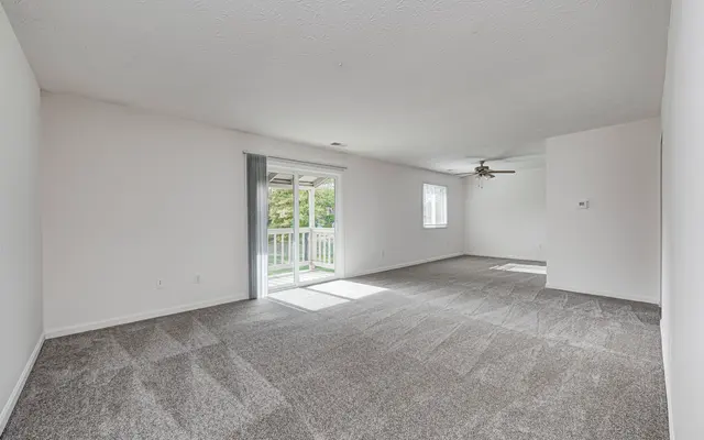 A spacious and bright living room with light gray carpet, white walls, and large windows that let in natural light. There is a ceiling fan and sliding doors leading to a balcony.