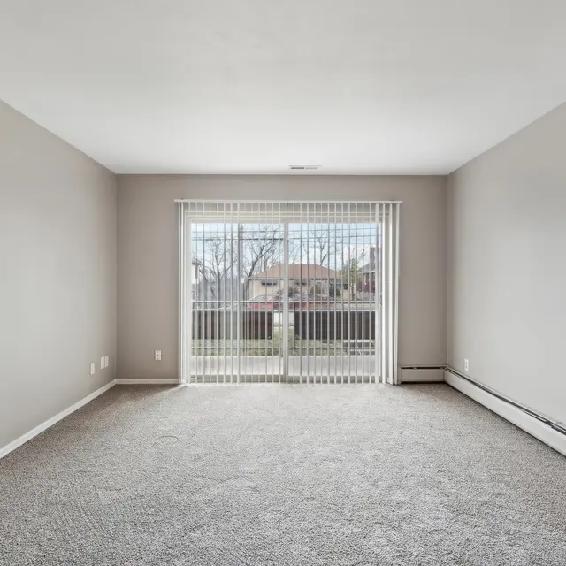 A spacious, empty living room with light gray walls, carpeted floor, and large sliding glass doors covered with vertical blinds that lead to a balcony.
