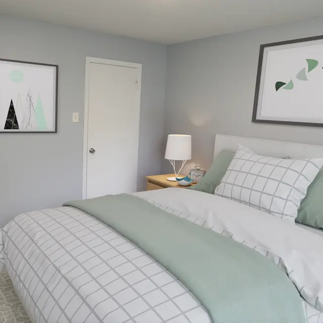 A modern bedroom featuring light gray walls, a neatly made bed with a mint green and white plaid comforter, and decorative artwork on the walls. A nightstand with a lamp and other items is present next to the bed.