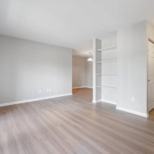 An empty living room with gray walls and light wooden flooring, featuring a built-in shelf and an adjacent kitchen area.