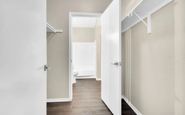 A view from a hallway into a bathroom and closet area, featuring beige walls and dark wood flooring.