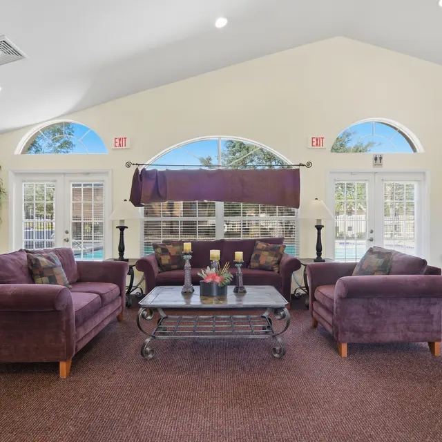A cozy lounge area featuring two purple sofas, a glass coffee table, and lush plants, with large windows allowing natural light.