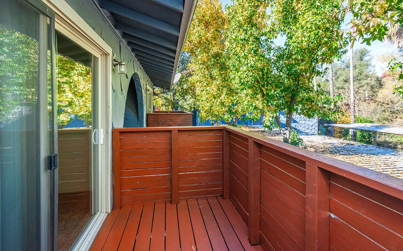 A wooden balcony with a brown railing overlooks greenery and trees. There are sliding glass doors leading into a room, and the area is illuminated by natural light.