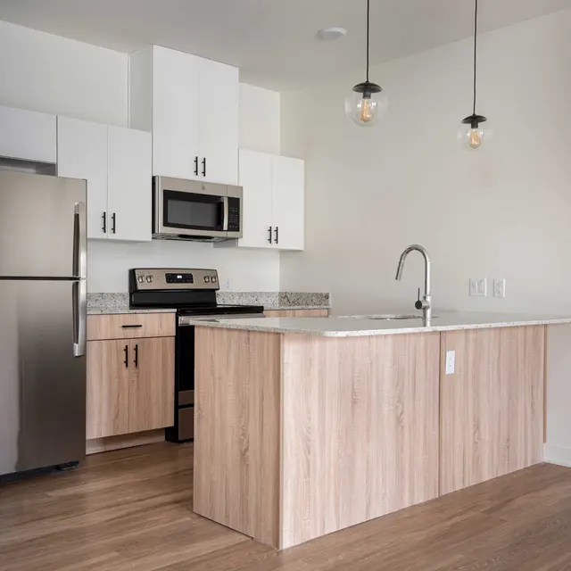 A modern kitchen featuring a stainless steel fridge, black stove and microwave, white upper cabinets, and light wood lower cabinets with a large island that has a white countertop.
