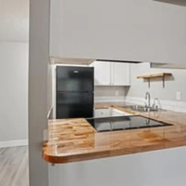 A modern kitchen featuring a wooden countertop, black refrigerator, and minimalist white cabinetry. Light gray walls and wooden shelves add to the contemporary look.