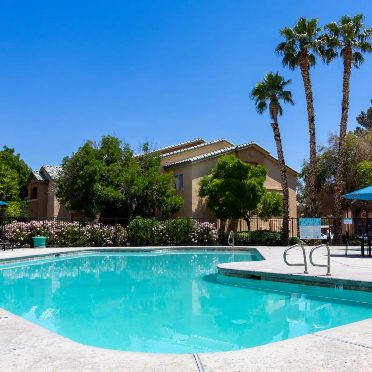 A tranquil swimming pool area featuring clear turquoise water, surrounded by lush green trees and palm trees. There are lounge chairs and umbrellas placed strategically around the pool, with a sunny blue sky in the background.