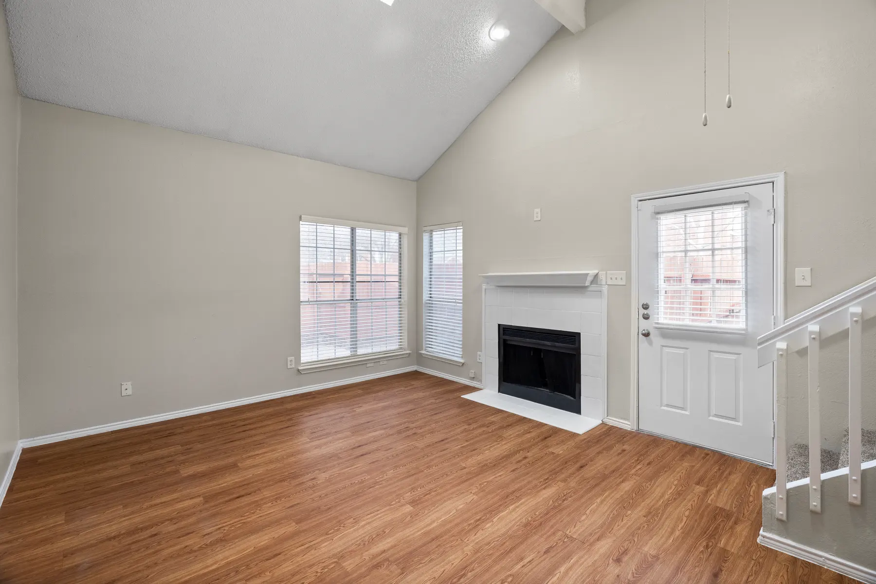 A spacious living room with high ceilings, large windows, and a fireplace. The floor is wooden, and there is a door leading outside as well as a staircase to the right.