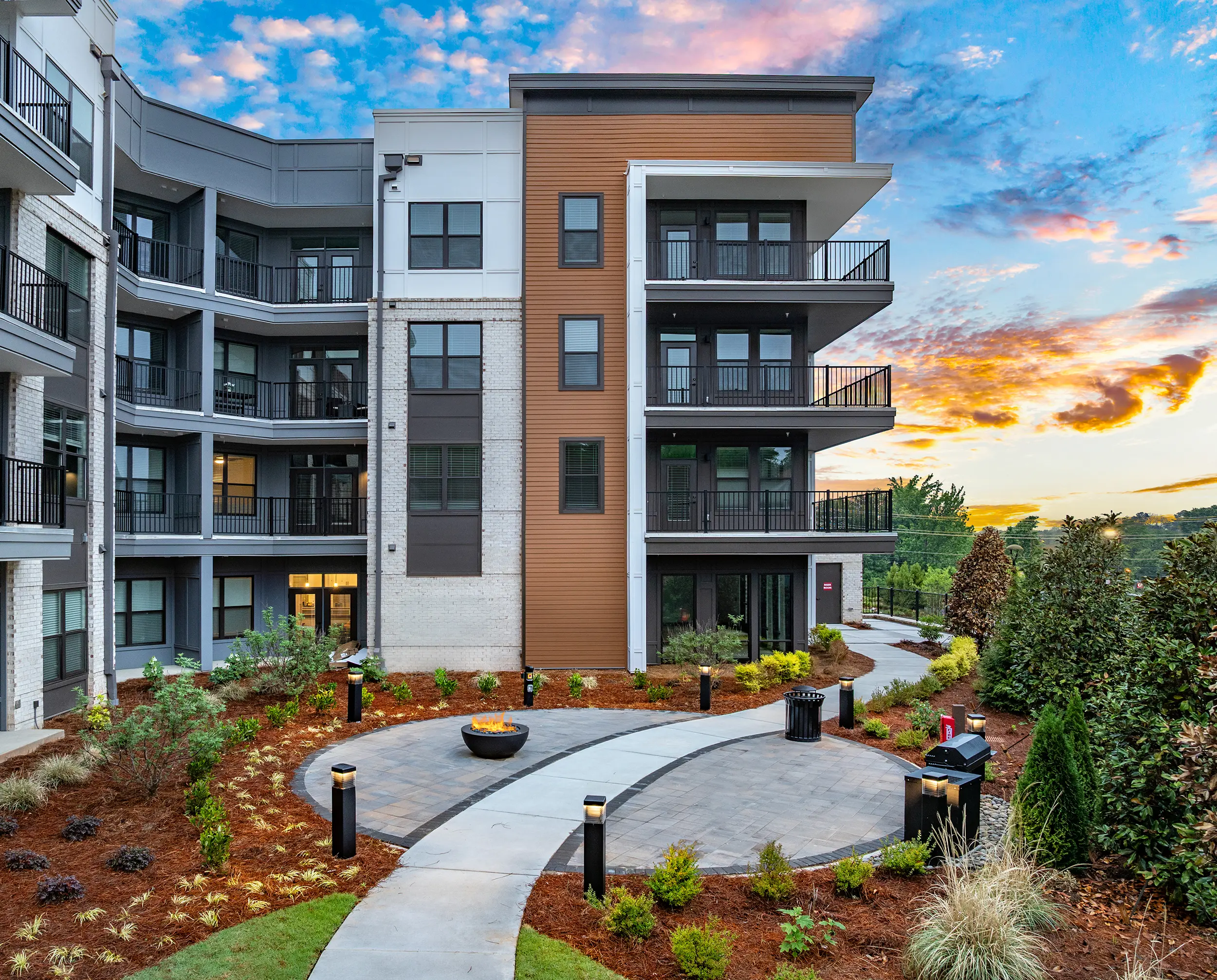 Modern Apartment Building at Sunset A modern apartment building with a combination of gray and brown exterior, featuring balconies and surrounded by landscaped greenery. The sky is colorful with clouds during sunset.
