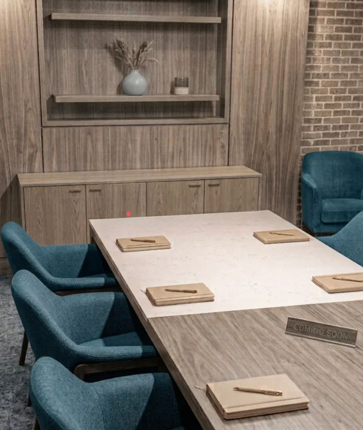 Modern Conference Room Design A modern conference room featuring a large table with pens and notepads placed on it, surrounded by blue chairs. A wooden shelf with decorative items is in the background.