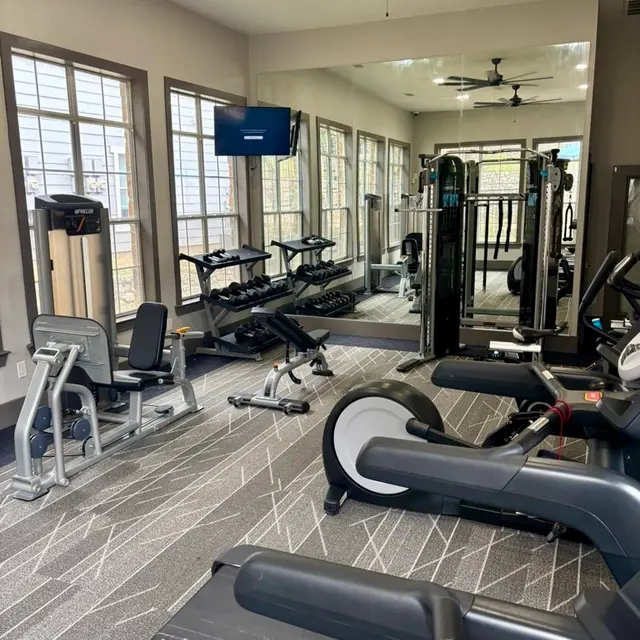 A spacious gym interior featuring various exercise equipment, including treadmills, weight machines, and free weights, with large windows and natural light.