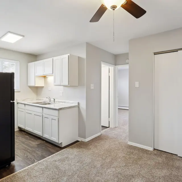 A modern kitchen with white cabinetry, wooden flooring, and a black refrigerator. There is a small area with carpet leading to a doorway.
