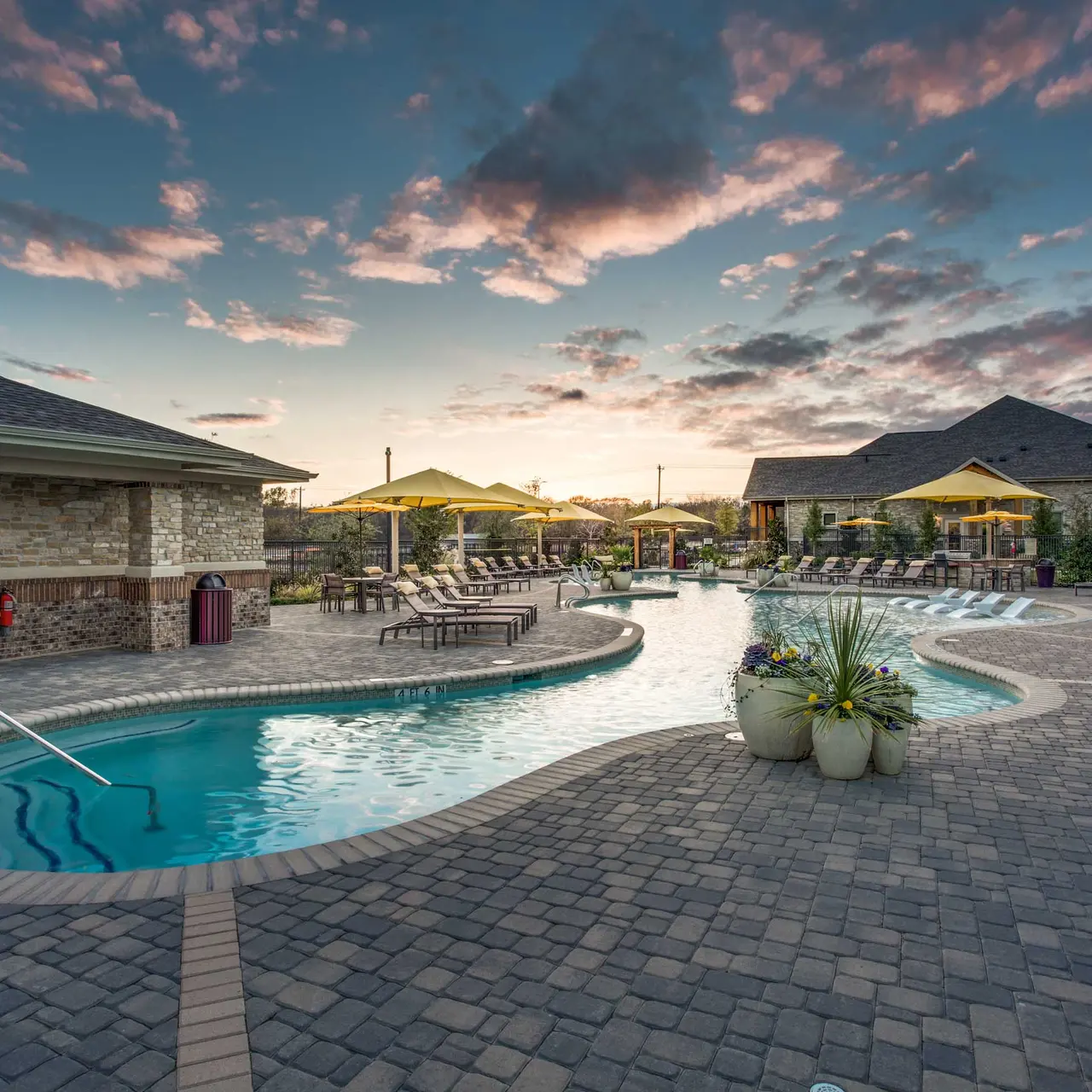 A serene pool area featuring a winding pool surrounded by lounge chairs and umbrellas, set against a sunset sky with colorful clouds.
