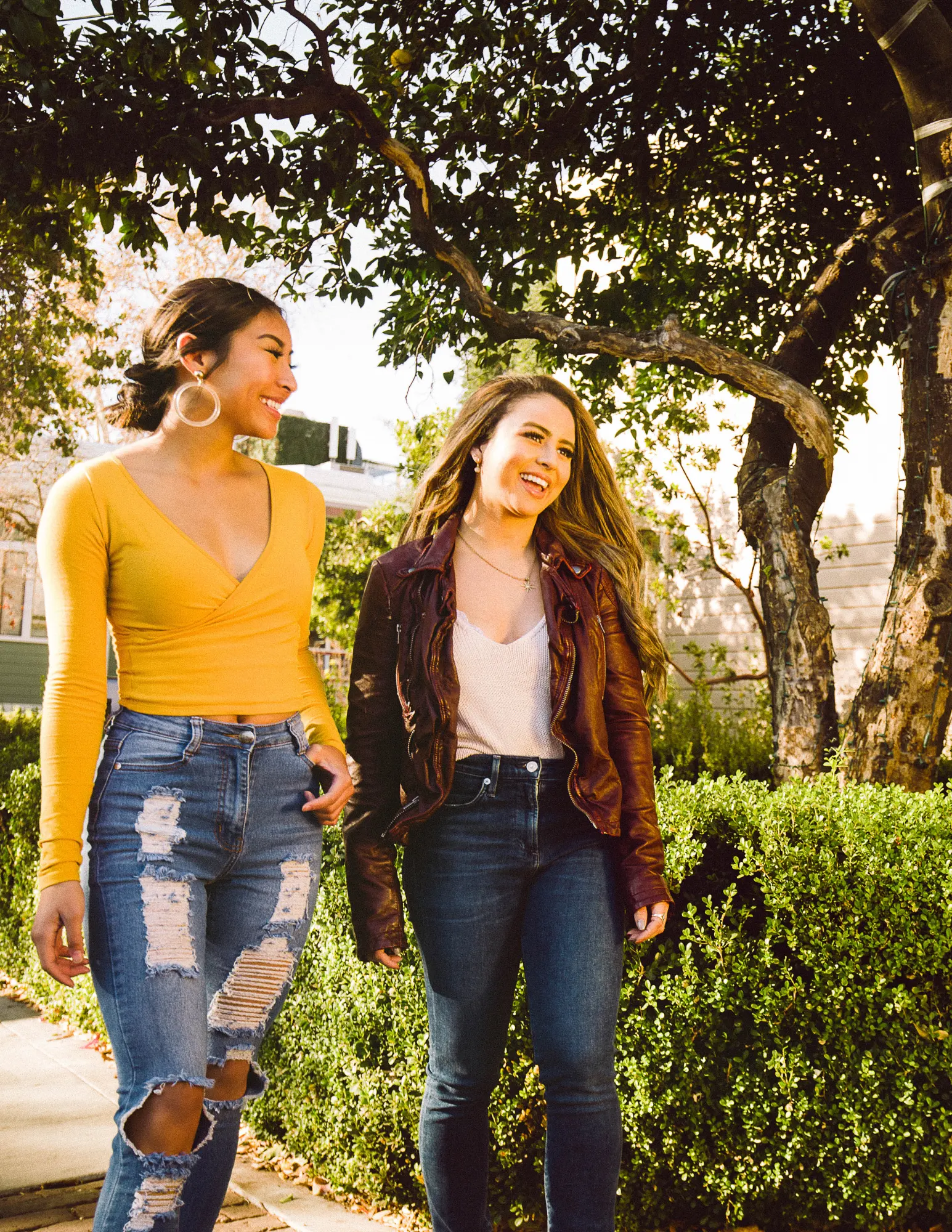 Two women walking and laughing in a garden setting. One wears a yellow long-sleeve top and ripped jeans, while the other wears a white top and a brown leather jacket over her shoulders. Both have long hair and are smiling, surrounded by greenery.