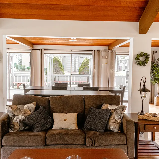 A cozy living room featuring a brown sofa with decorative pillows, two side tables with lanterns, and a dining area visible through large glass doors. Green plants and wreaths add natural touches.