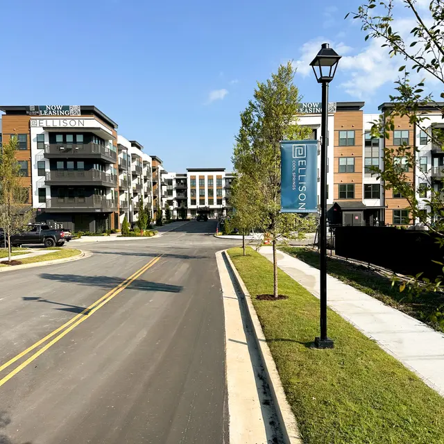 A wide street leading to a modern apartment complex on a sunny day. There are trees and lamp posts lining the street, with several vehicles parked along the side.