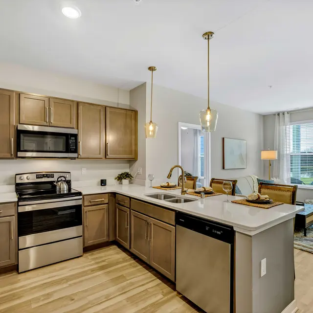 A modern kitchen featuring stainless steel appliances, wooden cabinets, and a spacious layout. The kitchen island has a double sink with a stylish faucet. The living area is visible in the background with natural light coming through large windows.
