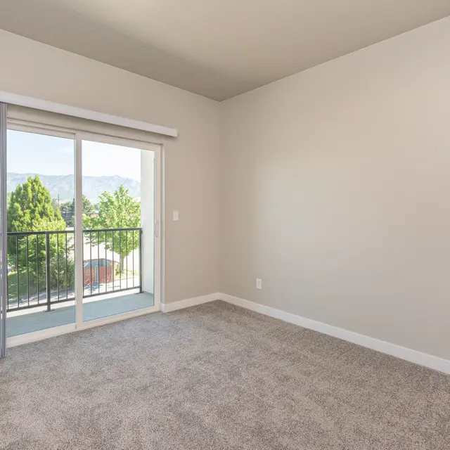 An empty room featuring beige walls, a doorway leading to a balcony, and carpeted flooring. Natural light is streaming in from the sliding glass door, showing greenery outside.