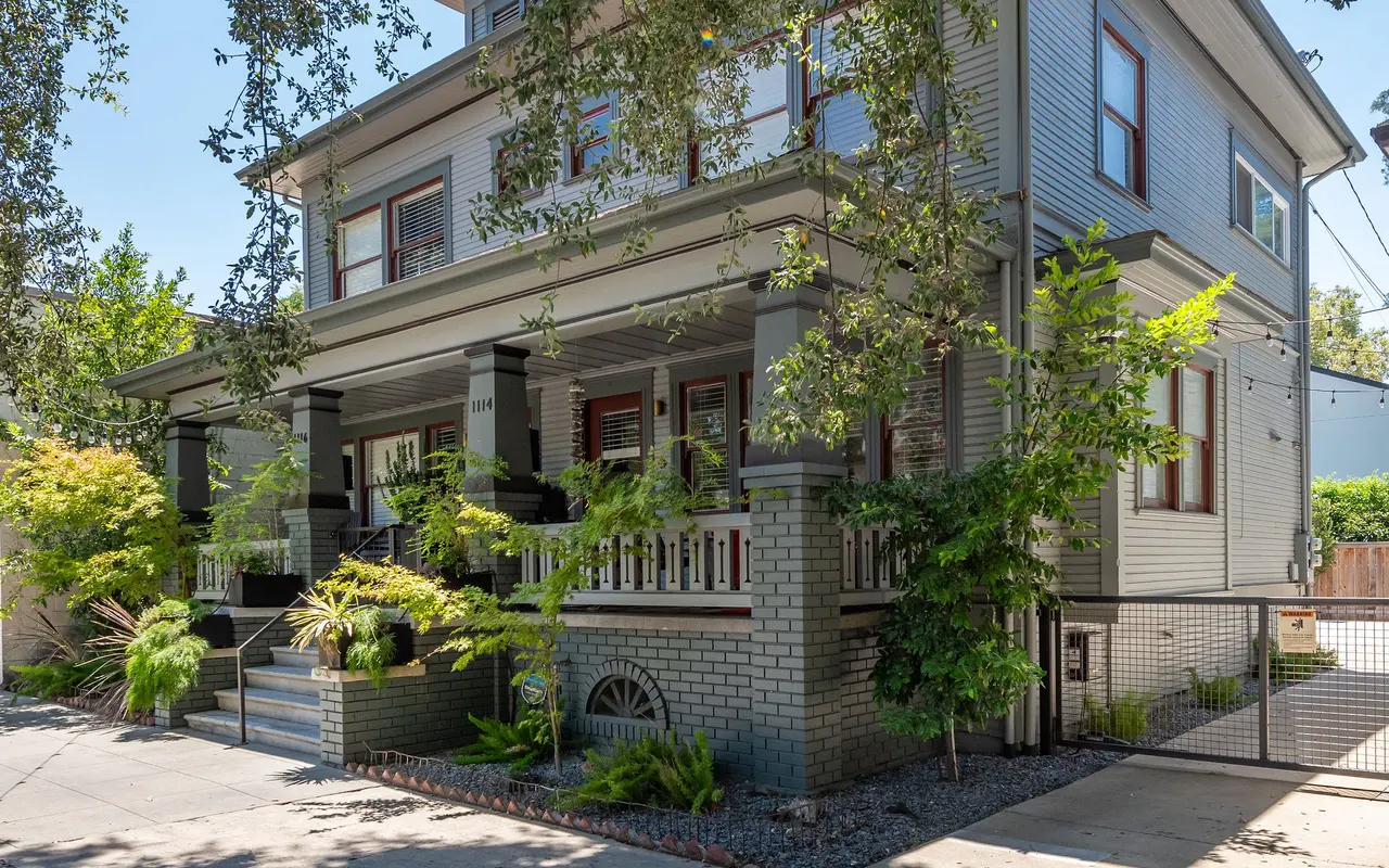 A Victorian-style house with a mix of gray siding and brick accents, surrounded by lush greenery. The entrance features a porch with steps leading up. Sunlight filters through the leaves of trees nearby.