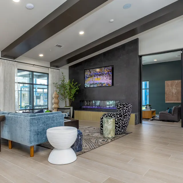 A modern living area with multiple seating options, including a plush blue couch and a white stool. There is a large table made of dark wood and a decorative rug on the floor. Large windows provide natural light, and a contemporary fireplace is mounted on the wall. A green plant adds a touch of greenery to the space.