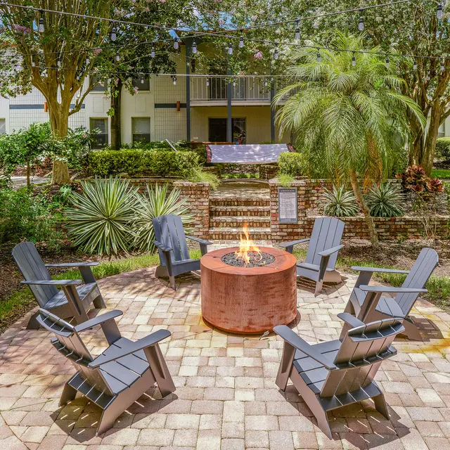 A circular fire pit surrounded by gray Adirondack chairs in a landscaped garden area, with lush plants and trees in the background.