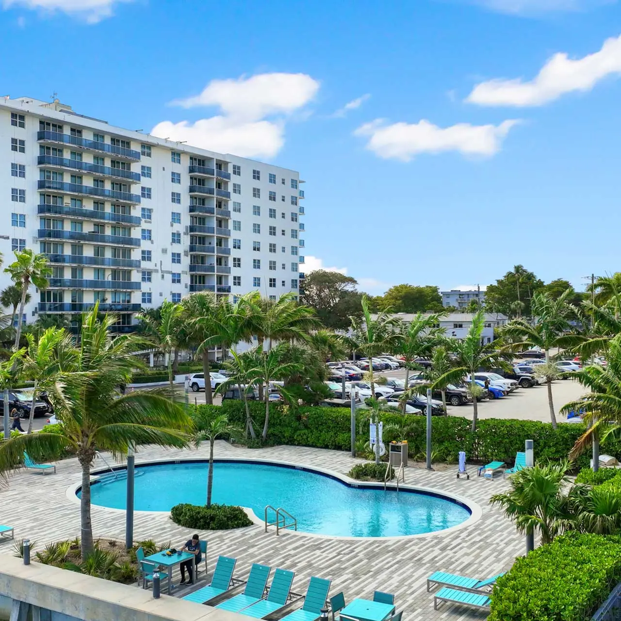 A view of a swimming pool surrounded by palm trees, with a large residential building in the background and parked cars visible nearby.