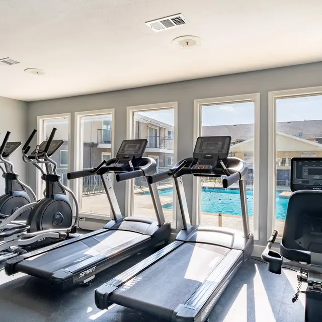 A modern gym interior featuring various exercise machines, including treadmills and ellipticals, with large windows overlooking an outdoor pool area.