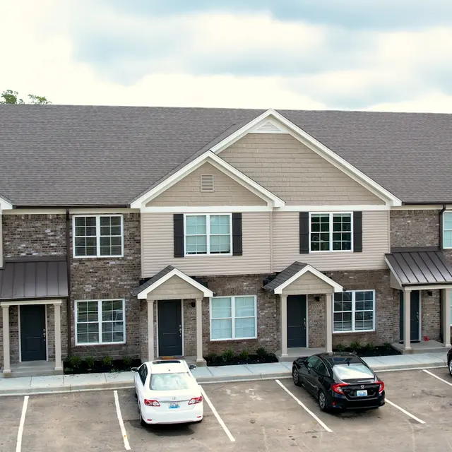A modern townhouse complex with multiple units featuring a mix of stone and siding architecture, surrounded by a parking lot with several cars parked.