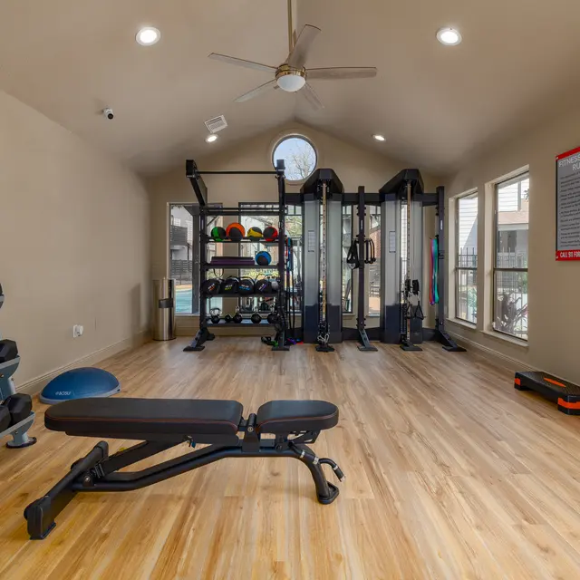 Interior view of a modern home gym featuring various exercise equipment, including dumbbells, a weight bench, and a multi-functional gym station, with wooden flooring and large windows allowing natural light.