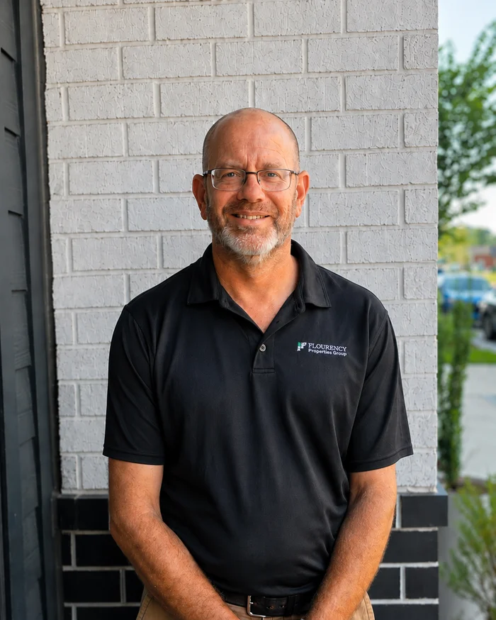 Portrait of a Smiling Man A smiling man wearing glasses and a black shirt stands outside against a brick wall. He appears friendly and approachable.