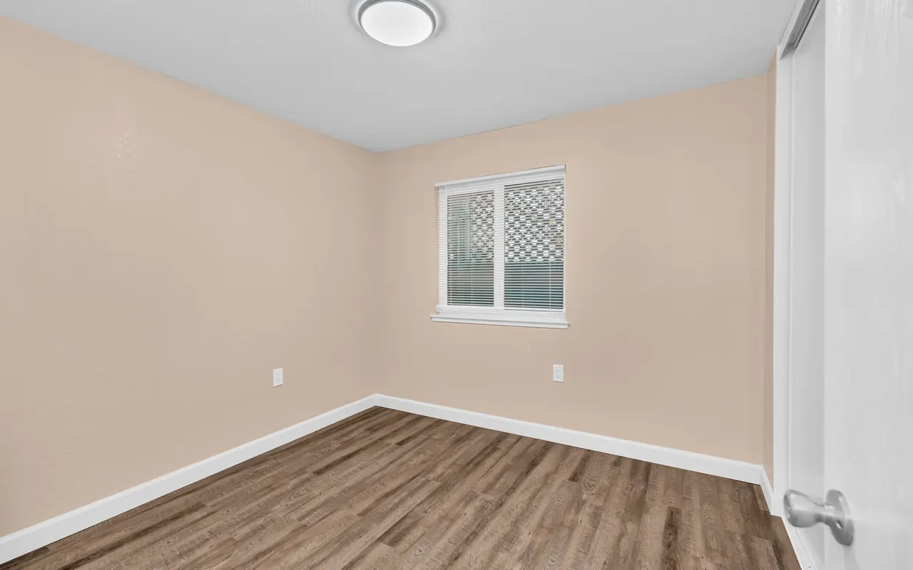 A vacant room with beige walls and a wooden floor, featuring a window with a patterned grill and a ceiling light fixture.