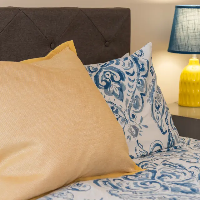 A cozy bedroom setting featuring a gray tufted headboard, decorative blue and white bedding, a large yellow pillow, and a yellow lamp with a blue shade on the nightstand.