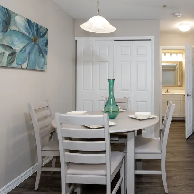 A dining area with a round table and four chairs, featuring a large flower painting on the wall. A green vase sits on the table, and there are plates set out. Light wood flooring is visible.