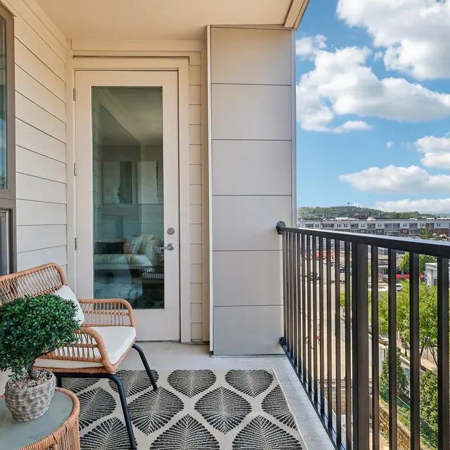 A balcony with a small seating area featuring a rattan chair, a round table with a potted plant, and a view of green trees and a clear blue sky with clouds in the background.