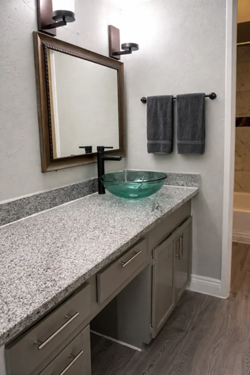 A modern bathroom with a gray granite countertop, featuring a round glass sink, a wall-mounted mirror, and two gray hand towels hung on a towel rack.