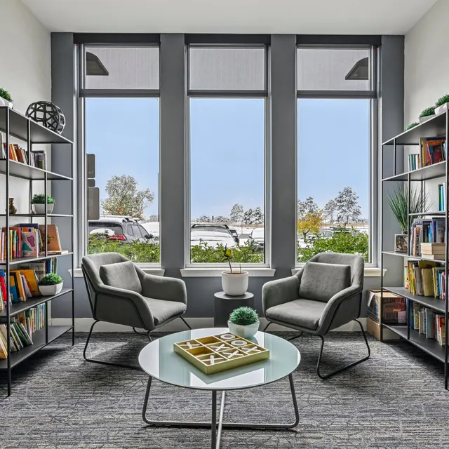 Cozy Reading Nook A cozy reading nook featuring two grey armchairs, a round glass table, and bookshelves filled with colorful books. Large windows allow natural light to fill the space, offering a view of greenery outside.