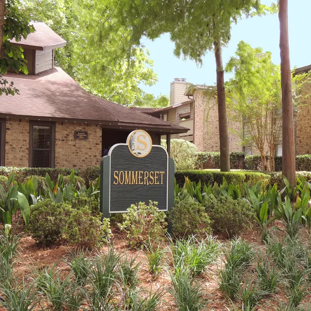 A sign for 'Somerset' located in a landscaped area with green plants and trees, next to a brick building under a blue sky.