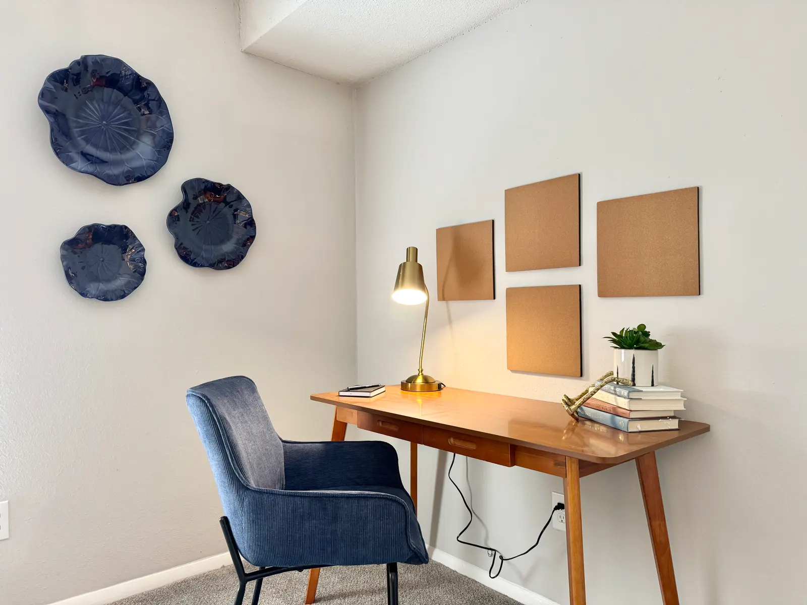A cozy modern home office featuring a wooden desk with books, a small potted plant, and a stylish lamp. A blue upholstered chair sits at the desk, while decorative plates adorn the wall. Four empty brown boards are mounted on the wall, creating a minimalist aesthetic.