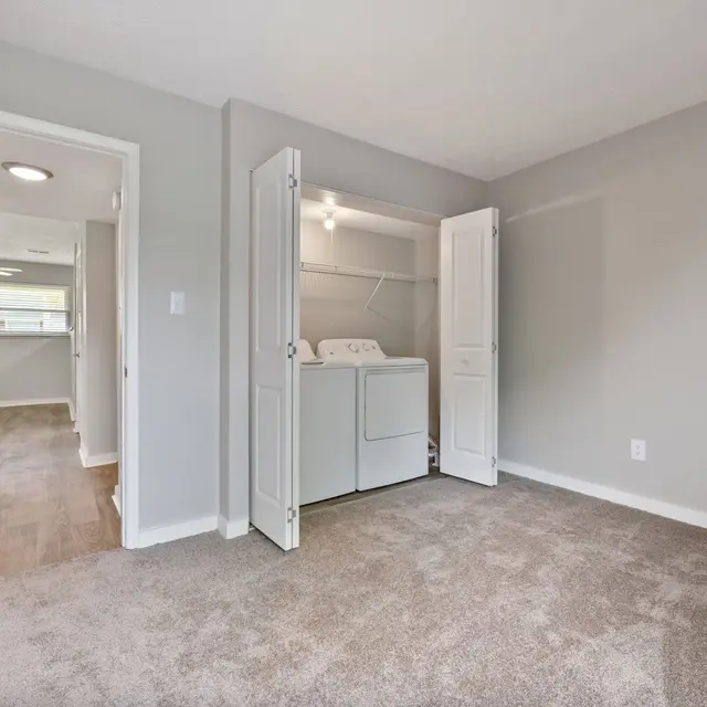 A clean, modern room with light gray walls, carpeted floor, and a laundry area visible in the open closet.
