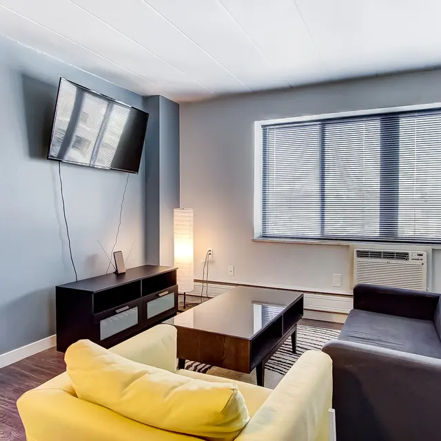 A modern living room featuring a black sofa, a yellow armchair, a dark coffee table, a TV mounted on the wall, large windows with blinds, and an air conditioning unit.