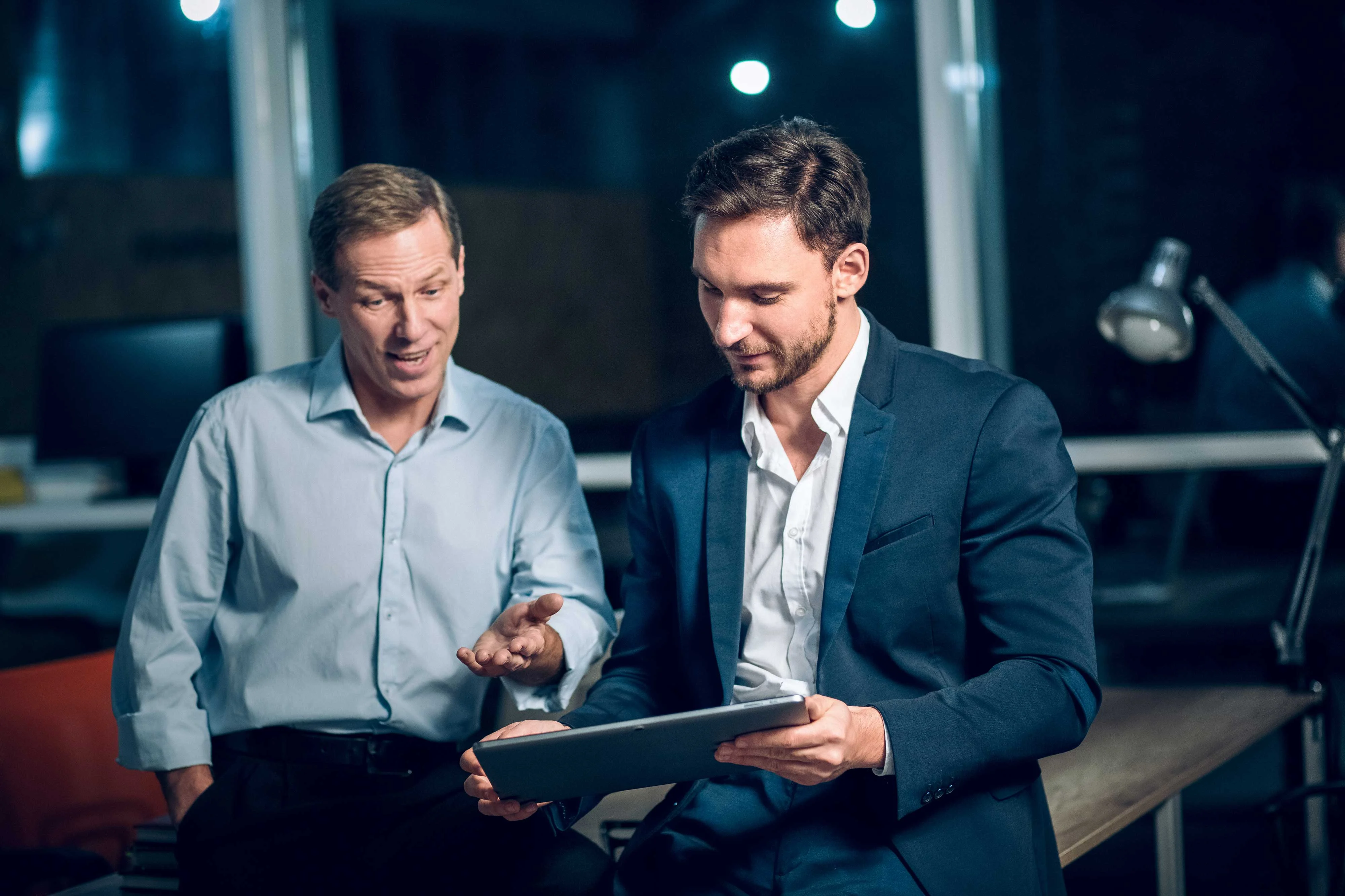 Evening Business Discussion Two men engaged in a discussion while looking at a tablet in a modern office setting.