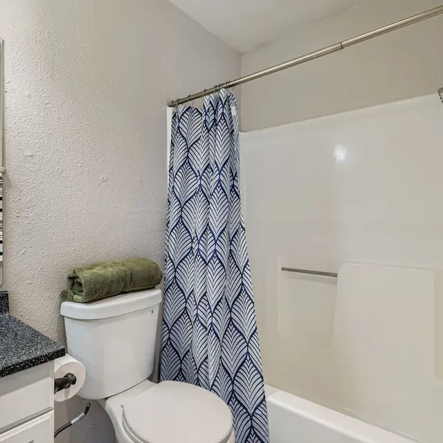 A small bathroom featuring a white shower curtain with a blue wave pattern, a black countertop sink with a mirror, and a toilet. A green towel is placed on the sink, and there are striped towels hanging nearby.