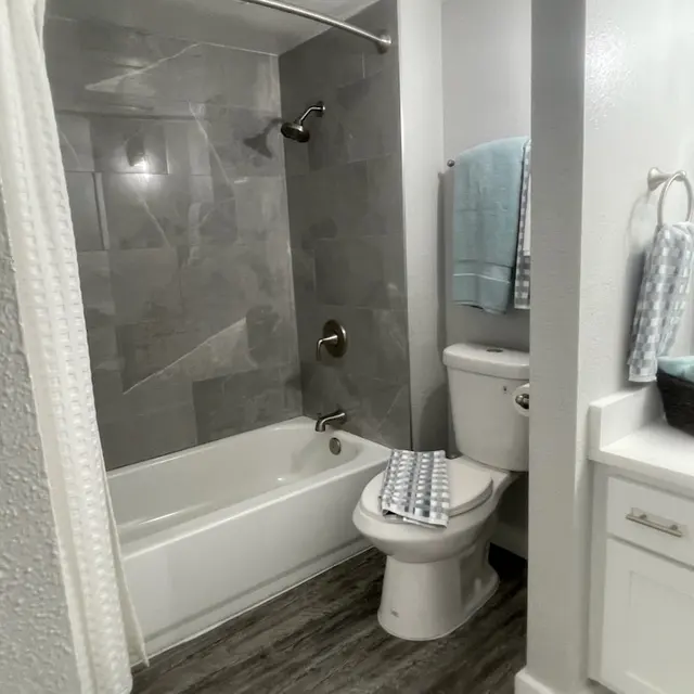 A modern bathroom featuring a bathtub with a shower, gray tiled walls, and light-colored flooring. A toilet is positioned to the side, and towels are hanging on a nearby rack.