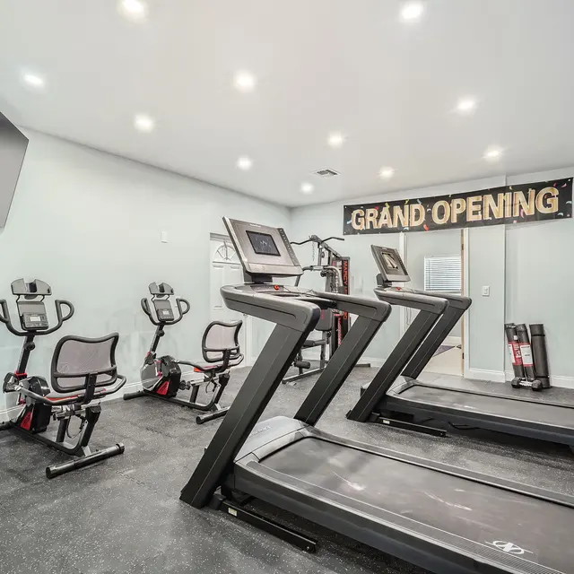 Interior of a gym featuring treadmills, exercise bikes, and a wall-mounted TV. A banner reads 'GRAND OPENING.'